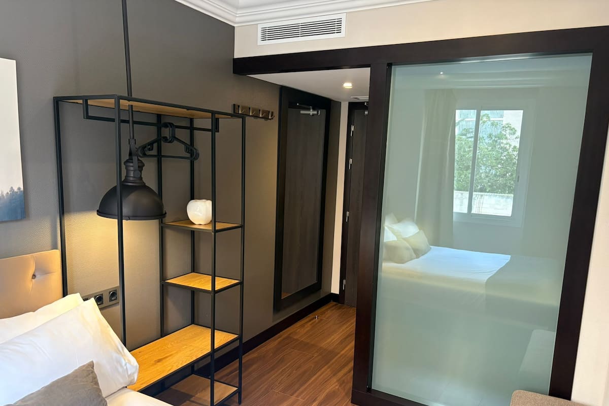 A modern hotel room with a bed, a desk, a chair, a lamp, and a sliding glass door leading to a balcony.