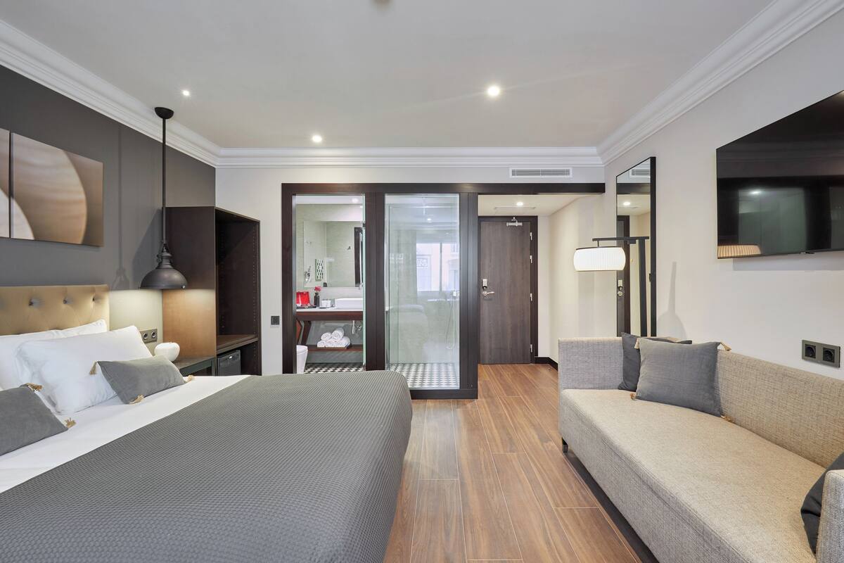 A modern hotel room with a large bed, a sofa, a TV, and a bathroom visible through a glass door.