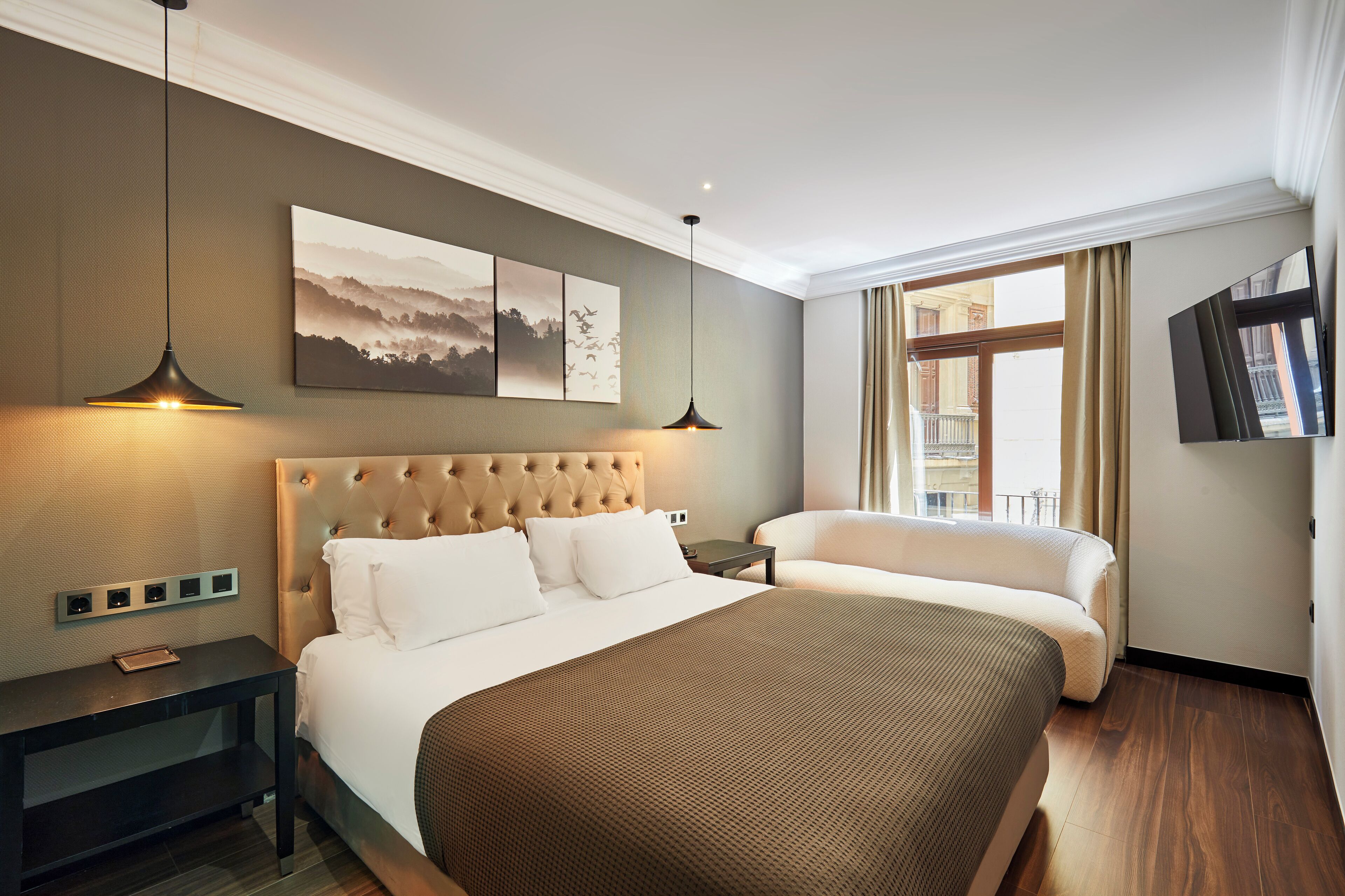 A hotel room with a large bed, a bedside table, a nightstand, a wall-mounted TV, and a window with curtains.