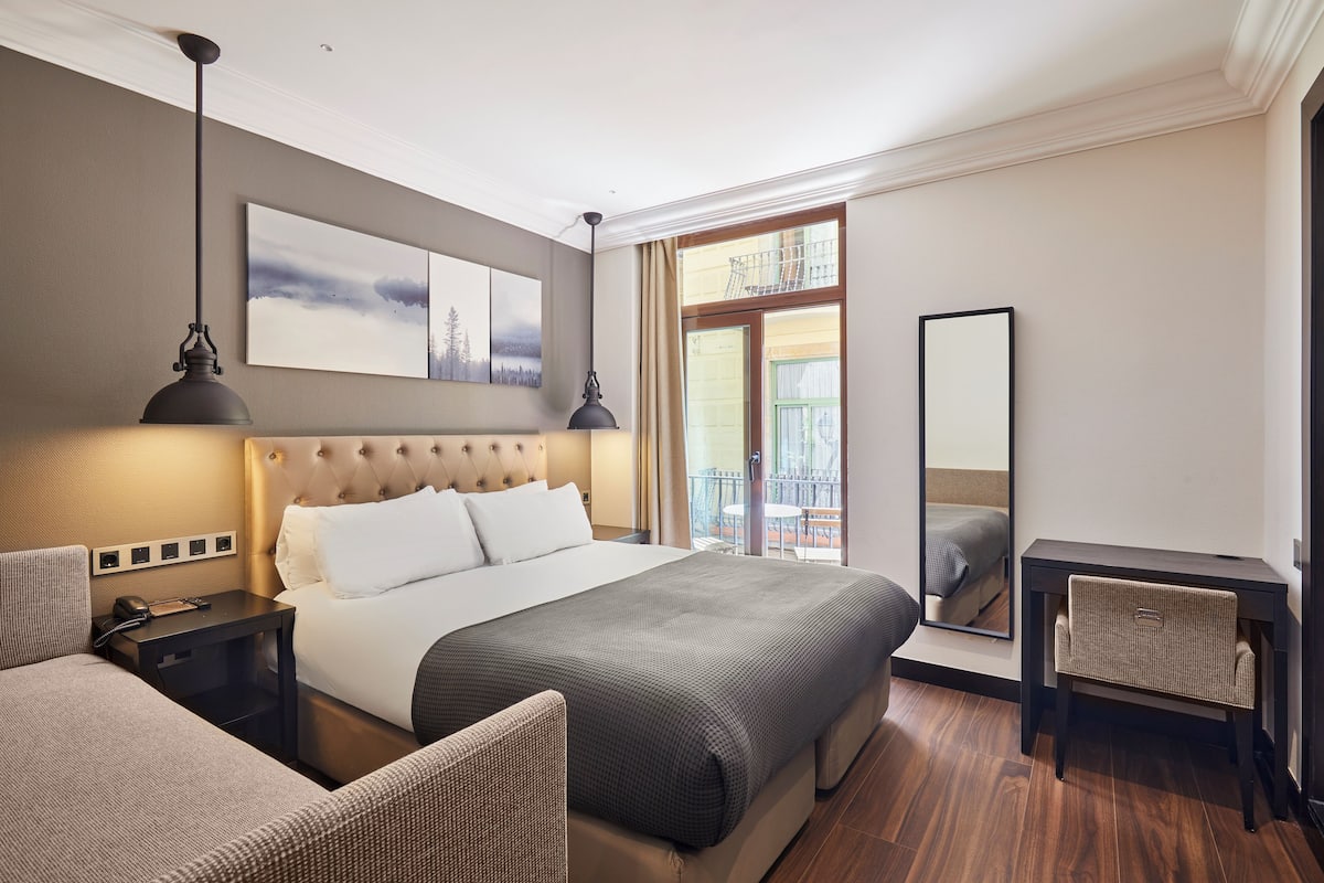 A hotel room with a large bed, a desk, a chair, a nightstand, a lamp, a painting, and a balcony view.