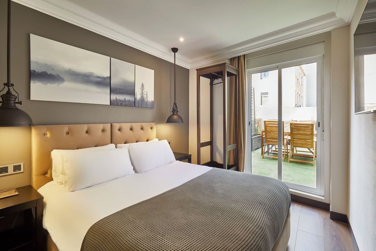 A hotel room with a large bed, a headboard, a nightstand, a lamp, and a sliding glass door leading to an outdoor area with chairs.