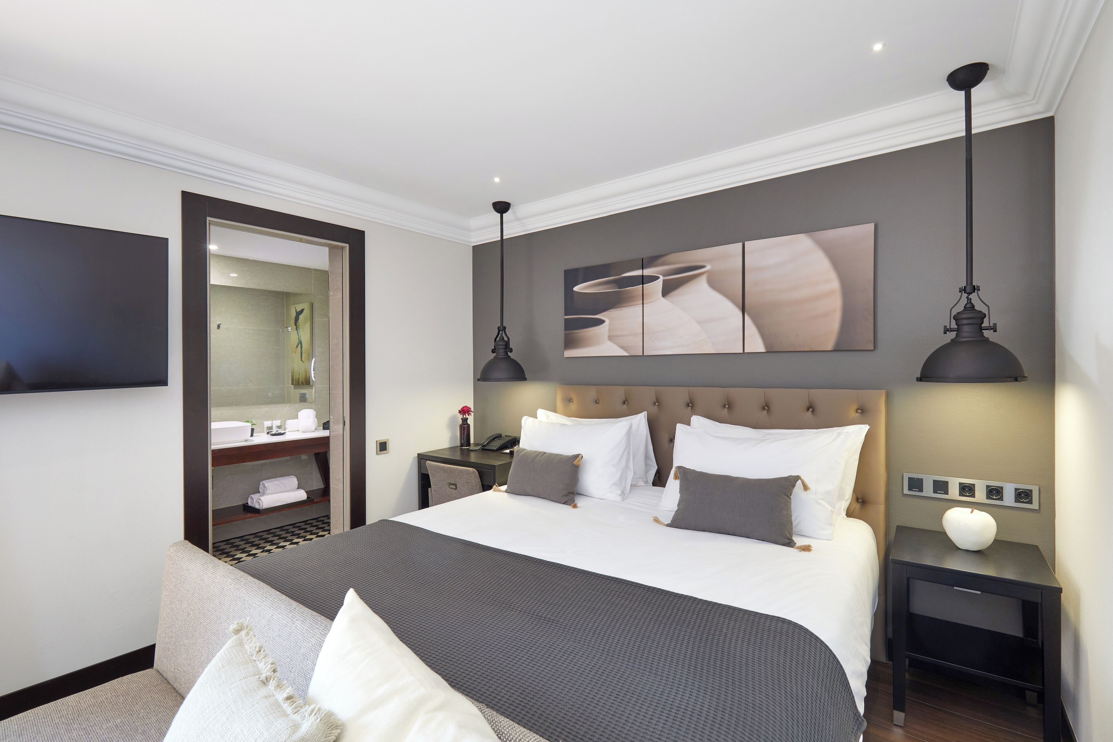 Ein modernes Hotelzimmer mit einem großen Bett, einem Nachttisch mit Lampe, einem zusätzlichen Hocker, einem am Wand montierten Fernseher und einem durchgehenden Türspalt sichtbaren Badezimmer.