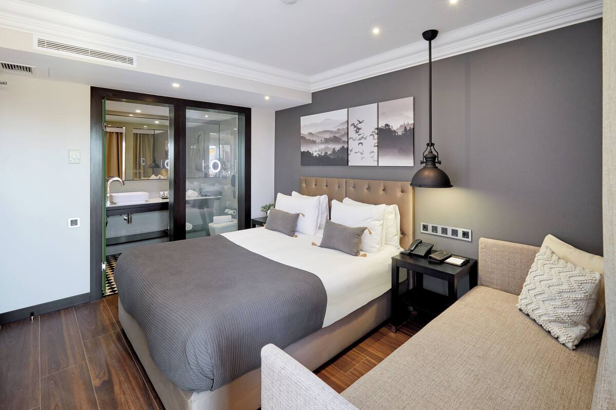 A modern hotel room with a large bed, a sofa, a desk with a lamp, and a bathroom visible through a glass door.