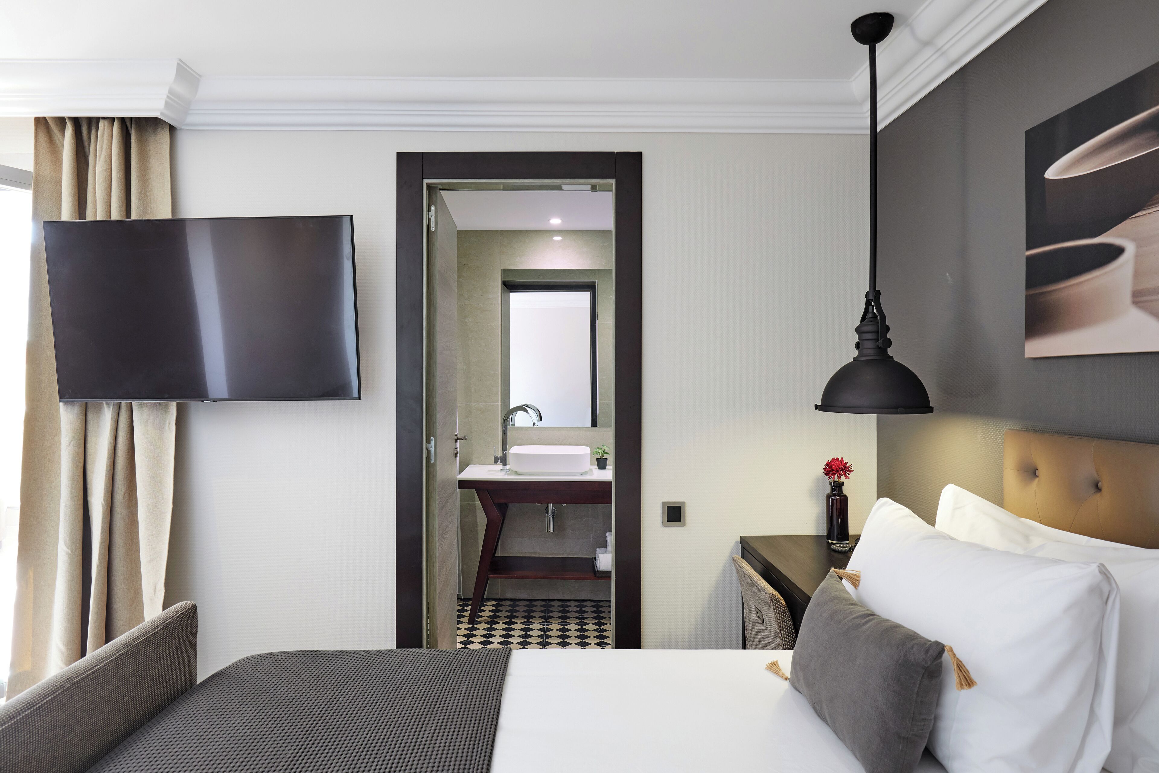 Habitación de hotel con cama, televisor de pantalla plana montado en la pared, mesita de noche con lámpara y baño visible a través de una puerta abierta.