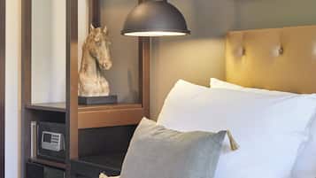 Una cama bien tendida con sábanas blancas y almohada gris, cabecera de madera, escultura de un caballo montado, lámpara de pared y un cuadro enmarcado sobre la cama.