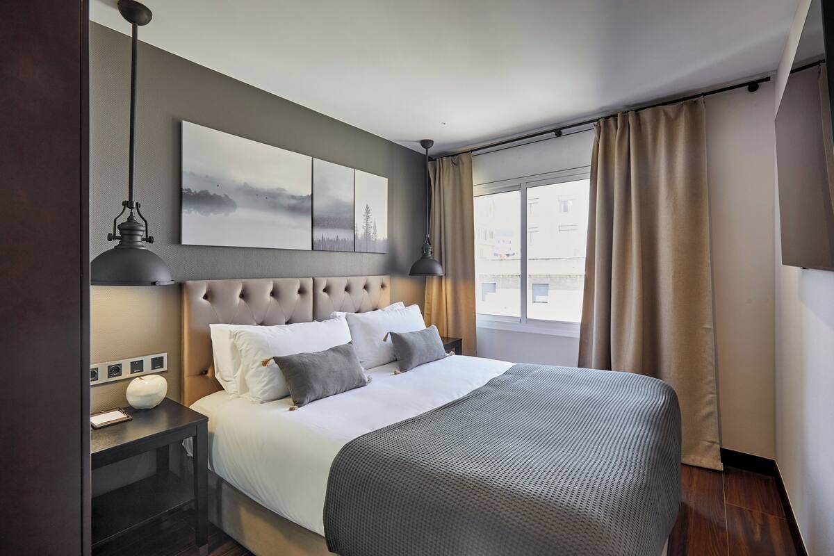 A hotel room with a large bed, a headboard, a bedside table with a lamp, a wall-mounted artwork, and a window with curtains.