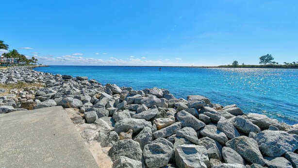 A rocky shoreline with a clear blue sea, a paved walkway, and distant buildings. - NEW BEACH LISTING, Walk-Beach, Surfing, Scuba, Close-Dining, Wi-Fi (West Palm Beach)