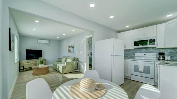 A modern living room with a sofa, a coffee table, and a dining area with a table and chairs. There is a kitchen area with white cabinets and appliances. - NEW BEACH LISTING-Block To The Beach, Fishing Nearby, Surfing, Chairs (West Palm Beach)