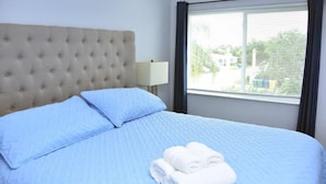 A hotel room with a tufted headboard, a large window with curtains, a bed with blue bedding, and a folded white towel on the bed. - NEW BEACH LISTING -Walk To Beach, BBQ Grill, Scuba, Beach Chairs (West Palm Beach)