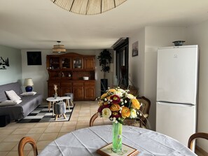 A living room with a sofa, a dining table with a vase of flowers, a kitchen area with cabinets, and a refrigerator.