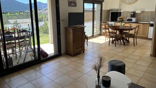 A spacious living area with a large sliding glass door leading to a balcony, a dining area with a wooden table and chairs, a kitchen with modern appliances, and a view of the mountains. - Le nid Capcinois (Formiguères)