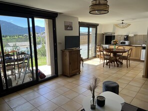 A spacious living area with a large sliding glass door leading to a balcony, a dining area with a wooden table and chairs, a kitchen with modern appliances, and a view of the mountains. - Le nid Capcinois (Formiguères)