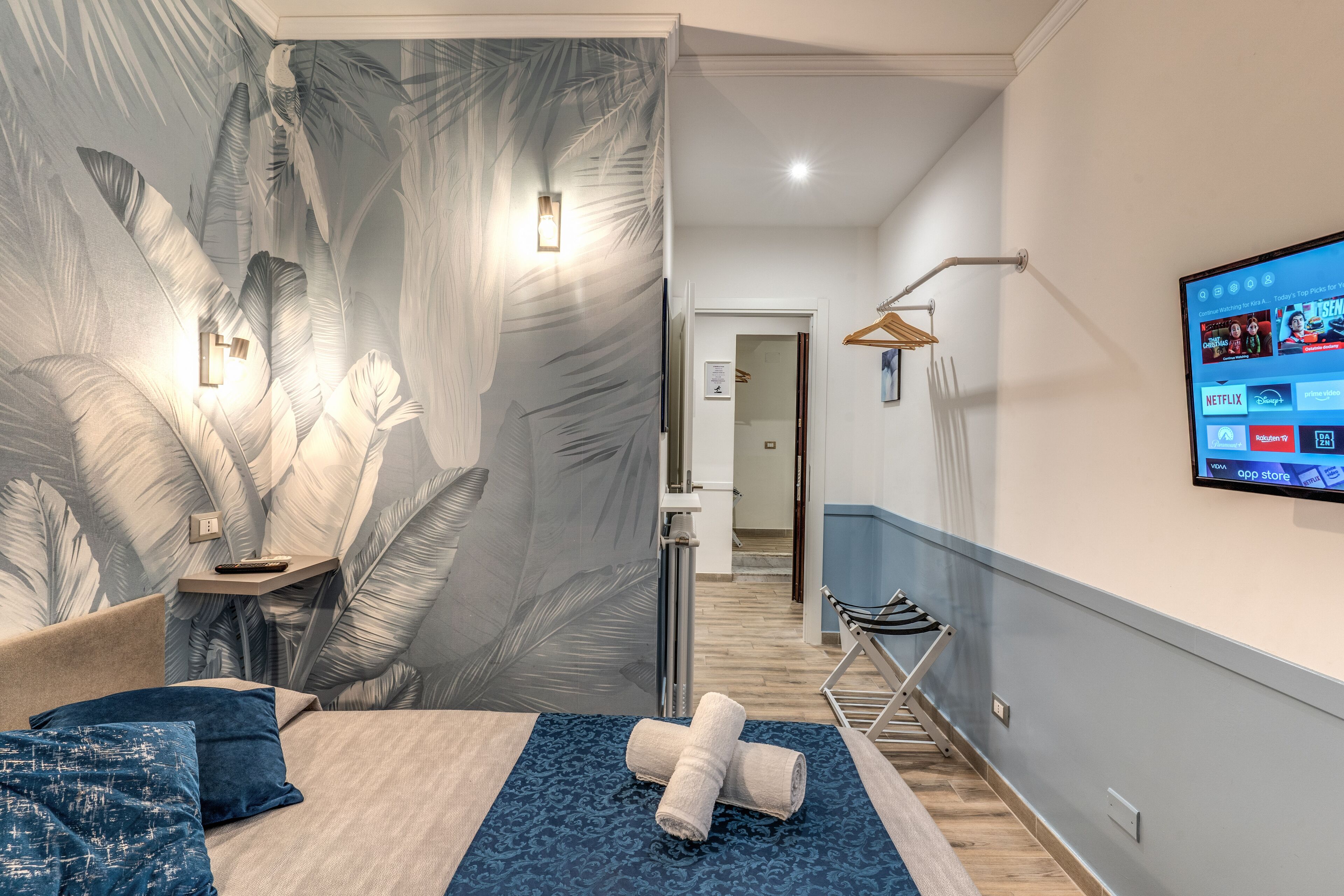 Une chambre d’hôtel moderne avec un grand lit, un téléviseur fixé au mur et un papier peint à motifs tropicaux.