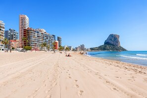 Uma extensa praia de areia com pegadas, céu azul límpido e uma formação rochosa imponente ao longe.