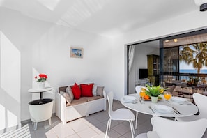 A modern living space with a dining area, a sofa with red cushions, a small table with a potted plant, and a view of the ocean through a glass door.
