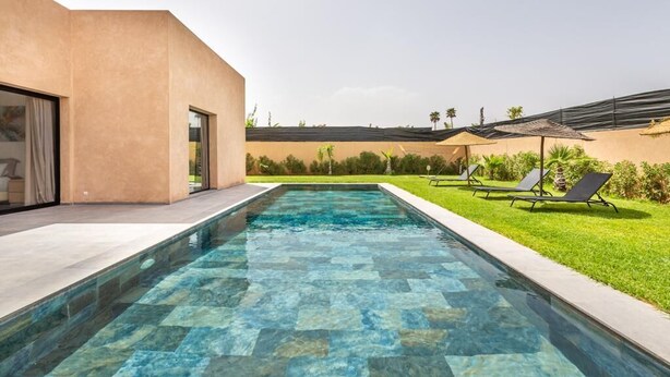 Una piscina rectangular con agua azul cristalina, rodeada por una solana empedrada. Hay sillones de descanso y un paraguas pequeño a la derecha.