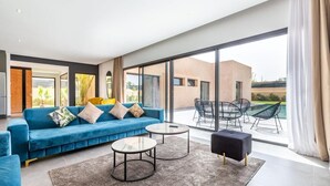 Una sala de estar moderna con un sofá azul, dos mesas de centro redondas y un reposapiés negro. Amplias ventanas ofrecen vistas al exterior.