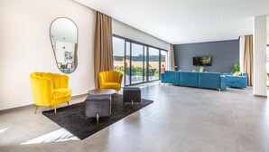 Un salón amplio con un ventanal grande, un espejo redondo, un sillón amarillo, un sofá azul y un televisor de pantalla plana.