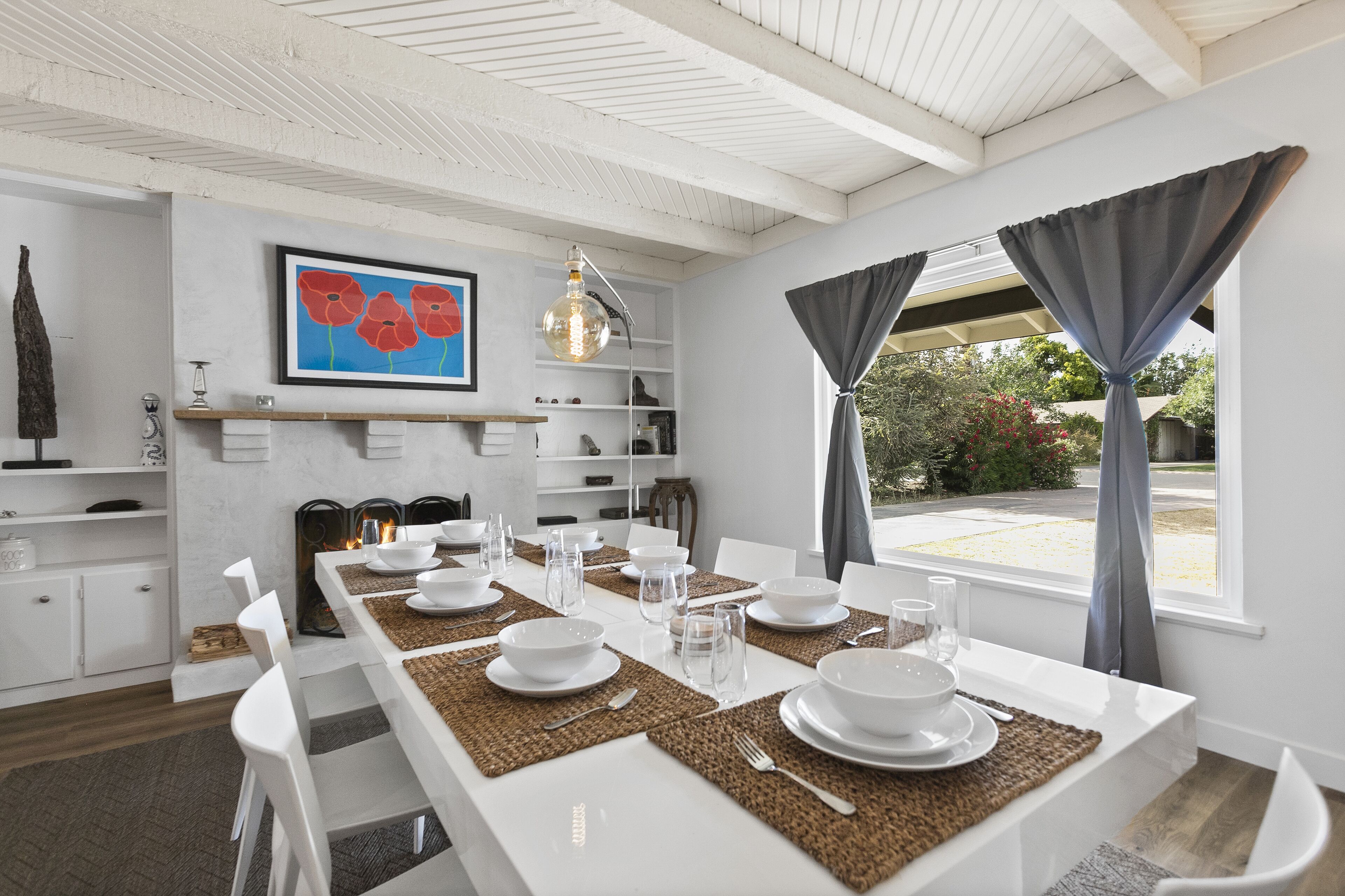 Uma sala de jantar com uma mesa comprida posta para refeição, cadeiras brancas, lareira e vista para o exterior através de grandes janelas.