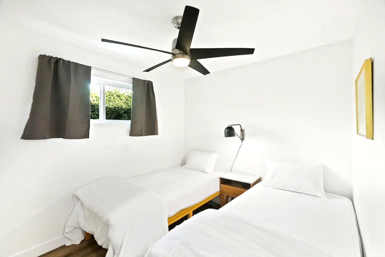 A room with two single beds, a ceiling fan, a bedside table with a lamp, and a window with curtains.