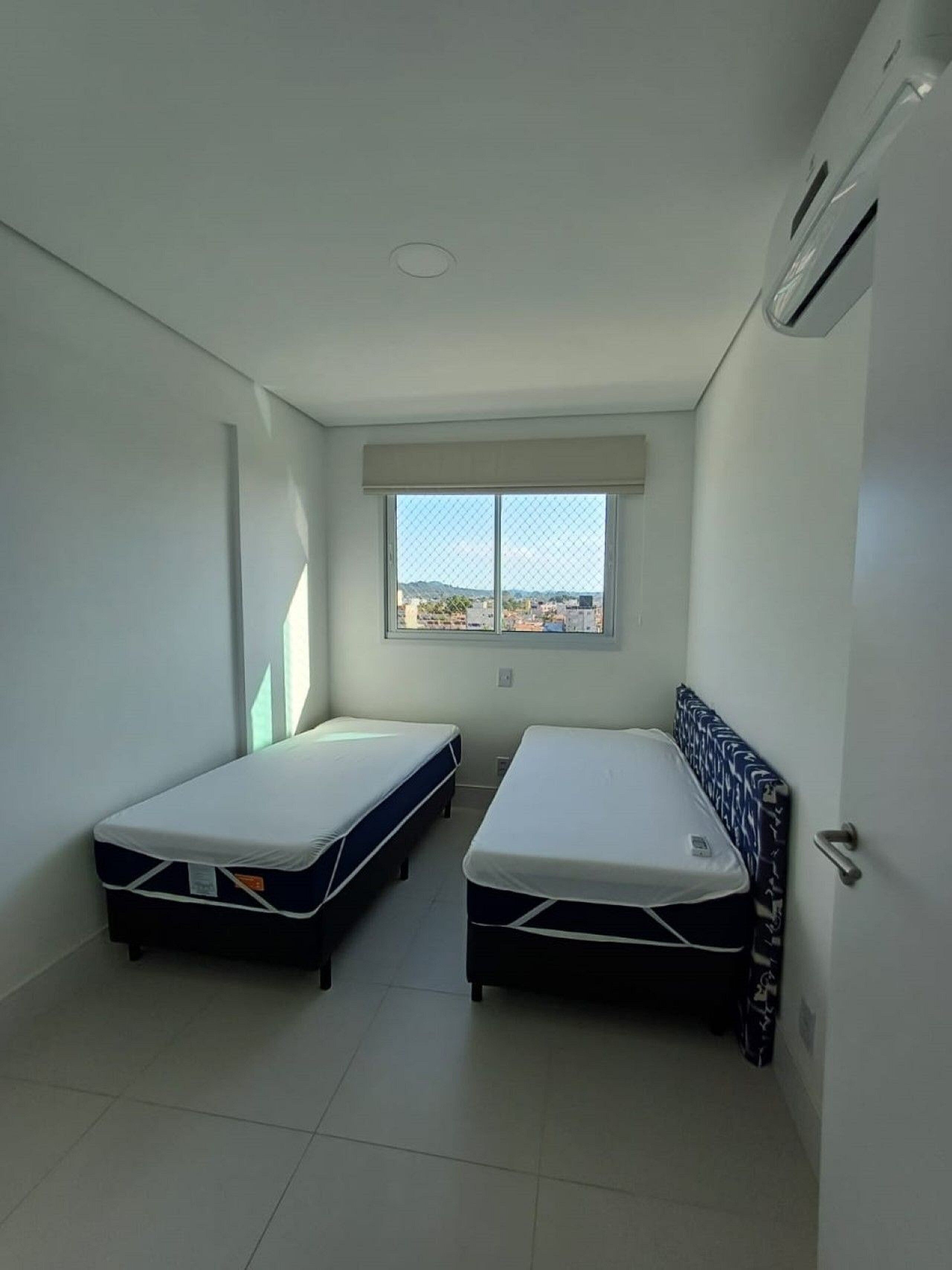 Een kleine, moderne kamer met twee enkele bedden, een groot raam met uitzicht op een stadsbeeld en een luchtgekoelde airconditioning.
