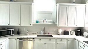 A kitchen with white cabinets, a microwave, a dishwasher, a sink, and a window with a blue curtain. - Beach Retreat Fun In The Sun Cozy Ambiance (Galveston)