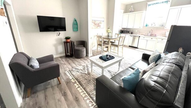 A modern living room with a sofa, armchair, and coffee table. There is a TV mounted on the wall and a dining area with a table and chairs. - Beach Retreat Fun In The Sun Cozy Ambiance (Galveston)