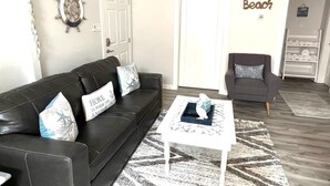 A living room with a black leather sofa, a white coffee table, and a gray armchair. There is a wall clock and a sign that says "Beach". - Beach Retreat Fun In The Sun Cozy Ambiance (Galveston)