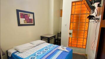Una habitación pequeña y funcional con cama, silla, mesa y ventana con reja de metal.