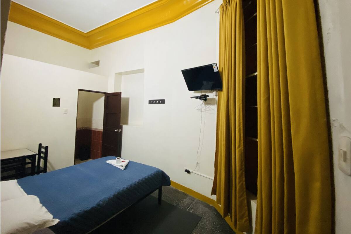 A hotel room with a bed, a desk, a chair, a television mounted on the wall, and a window with curtains.