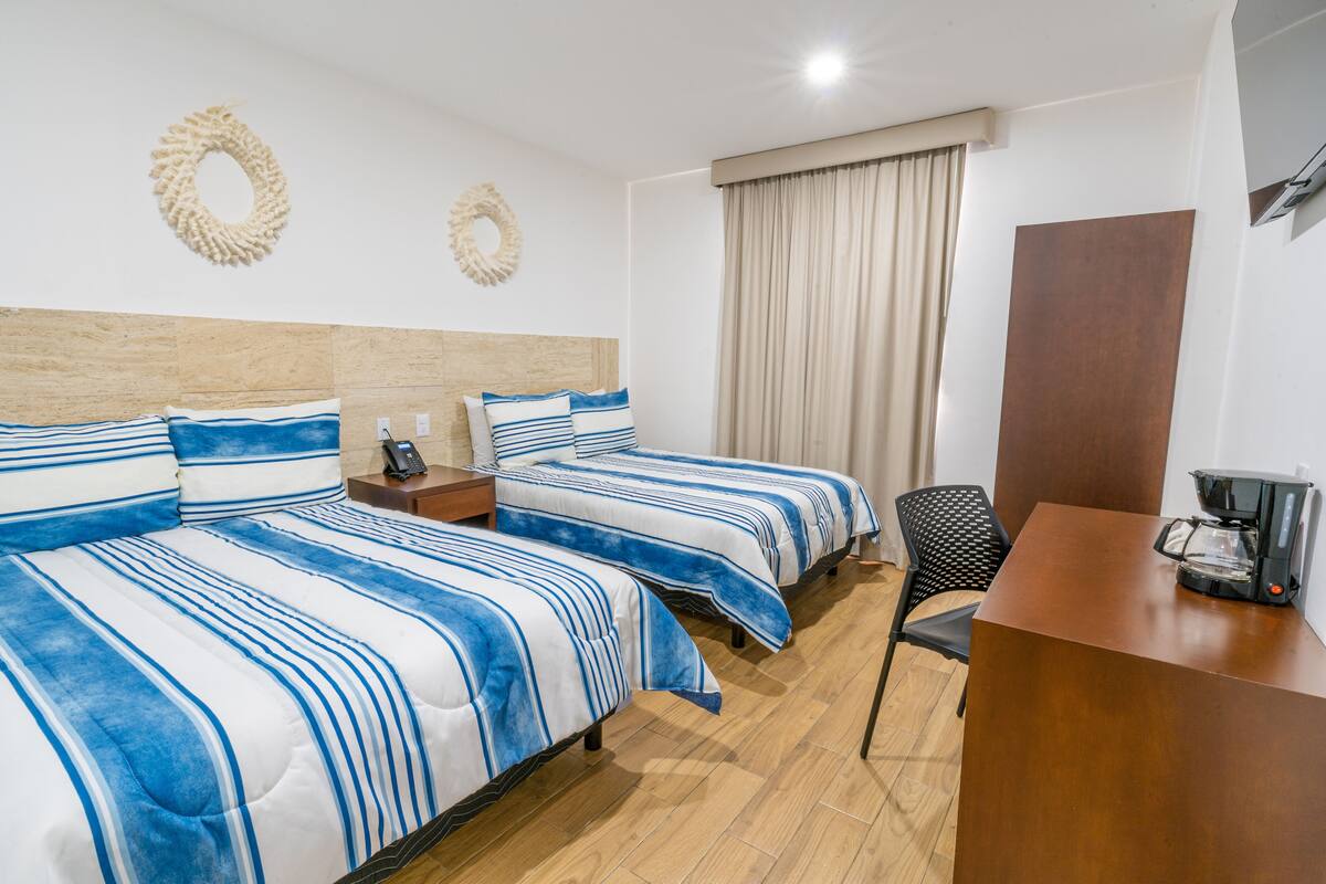 Quarto de hotel com duas camas, uma escrivaninha, uma cadeira, cafeteira e cortina.