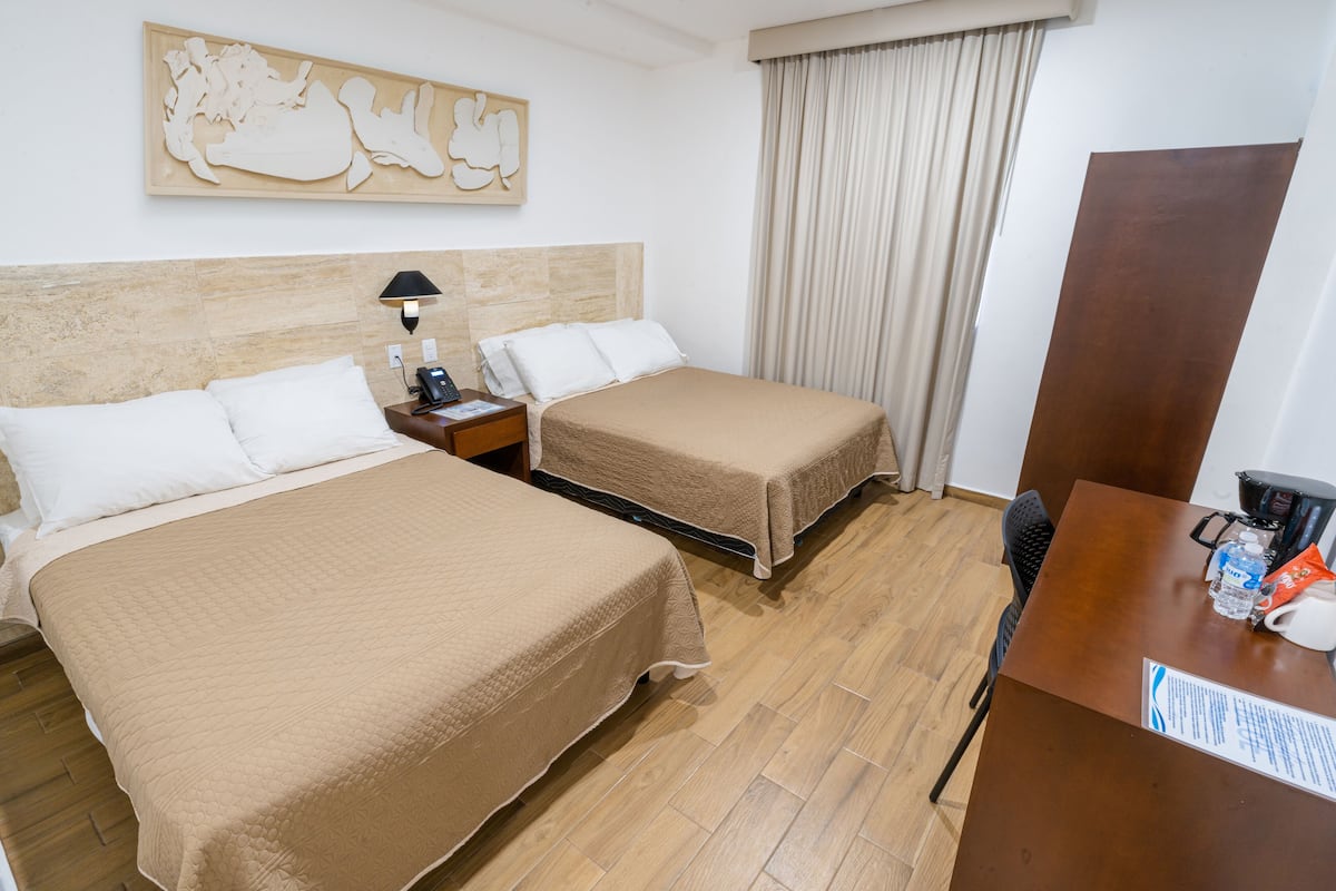 Quarto de hotel com duas camas, piso de madeira, uma escrivaninha com cadeira, máquina de café e uma obra de arte na parede.