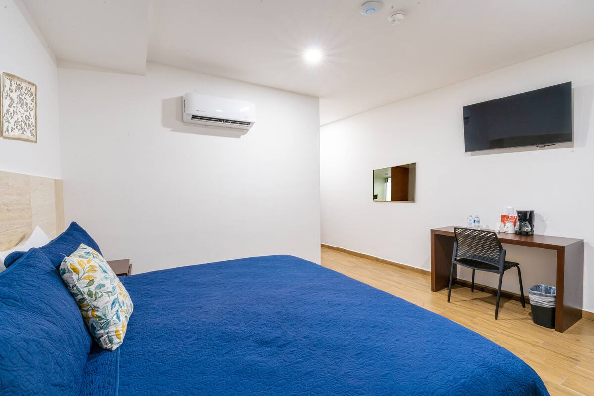 Quarto de hotel com uma cama grande, uma escrivaninha com cadeira, uma TV e um aparelho de ar condicionado.