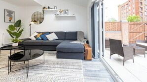 Un saló modern amb un sofà modular, un mirall rodó i una taula de centre. Hi ha una finestra gran que ofereix vistes a un edifici de maó i una tanca de fusta.