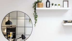 Un mirall rodó amb un dibuix de rejilla, una làmpada de taula negra, una estanteria blanca amb plantes i llibres, i una planta penjant amb fulles verdes.
