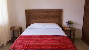 Een enkel bed met een houten hoofdeinde, wit beddengoed en een rode gebloemde deken. Twee houten nachtkastjes, waarvan er één een boek en een kleine vaas met bloemen heeft.
