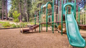 A playground with a slide and climbing structures, surrounded by trees. - Ski-in/Ski-out Northstar Condo (Truckee)