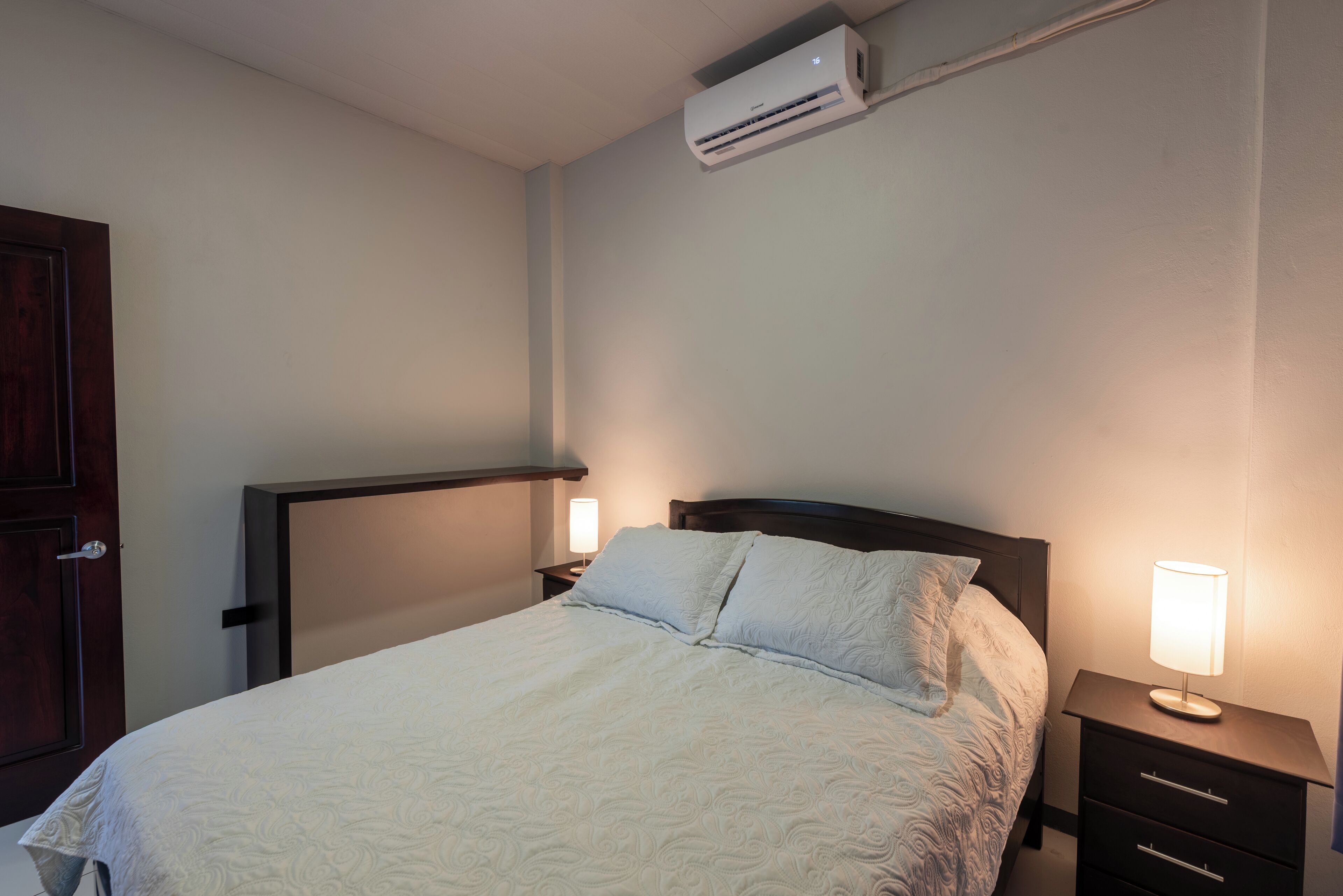 A neatly made bed with a white comforter and pillows, a bedside lamp, a dark wooden nightstand, and a wall-mounted air conditioner.