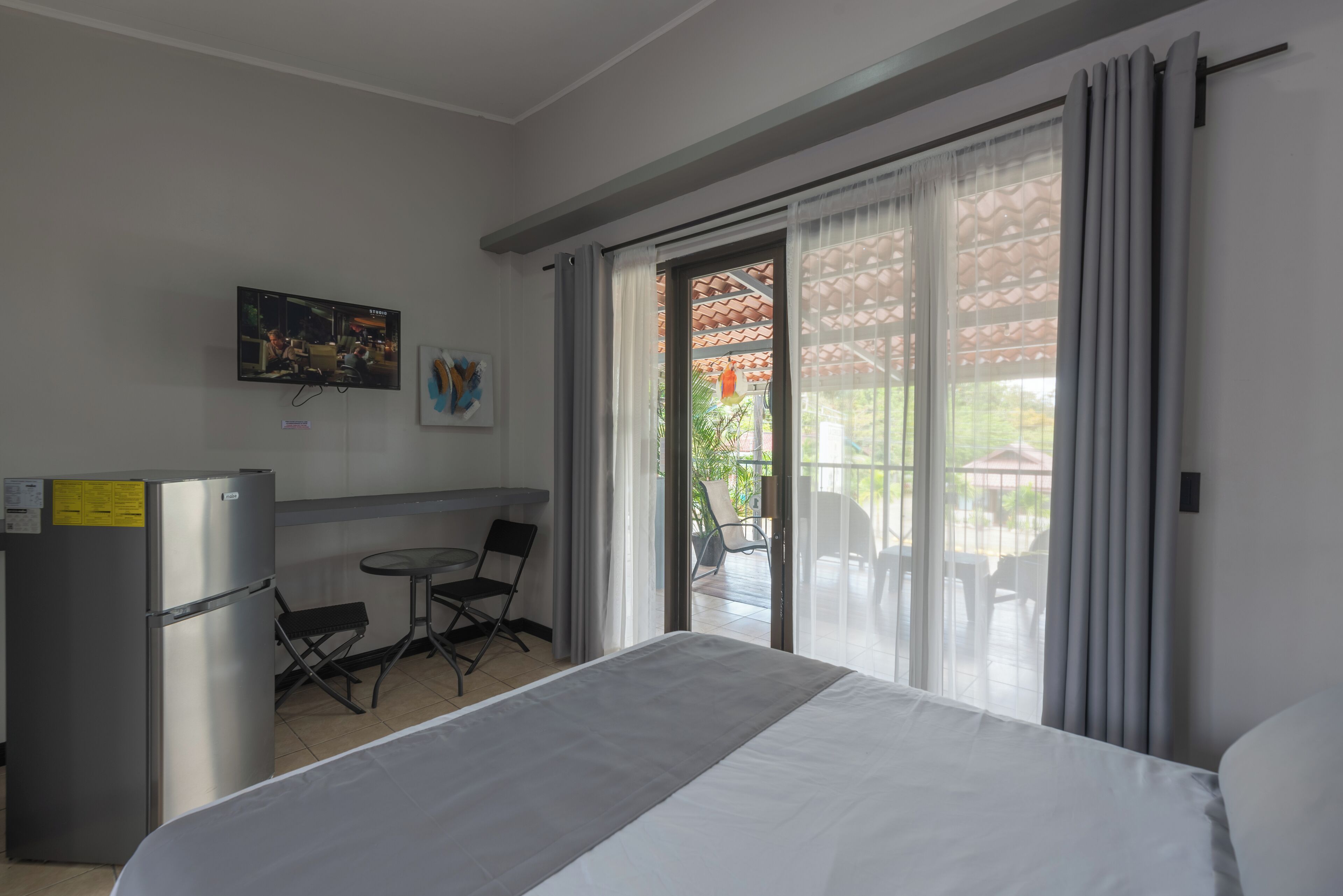 A hotel room with a bed, a small table, a chair, a refrigerator, and a sliding glass door leading to an outdoor area with chairs and a table.