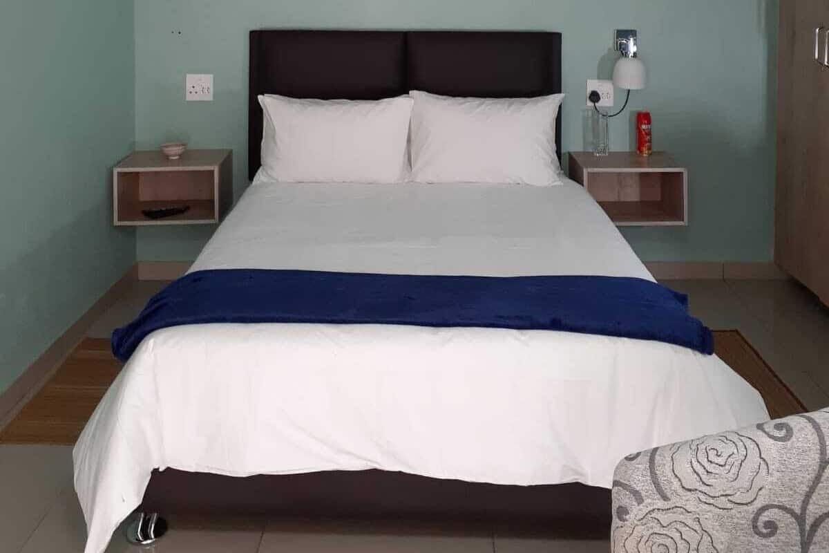 A bed with white bedding and a blue throw, flanked by two bedside tables with lamps and a small shelf, in a room with teal walls and wooden flooring.