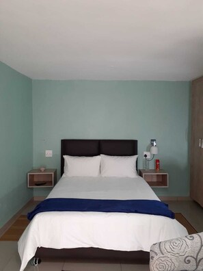 A bed with white bedding and a blue throw, flanked by two bedside tables with lamps and a small shelf, in a room with teal walls and wooden flooring.