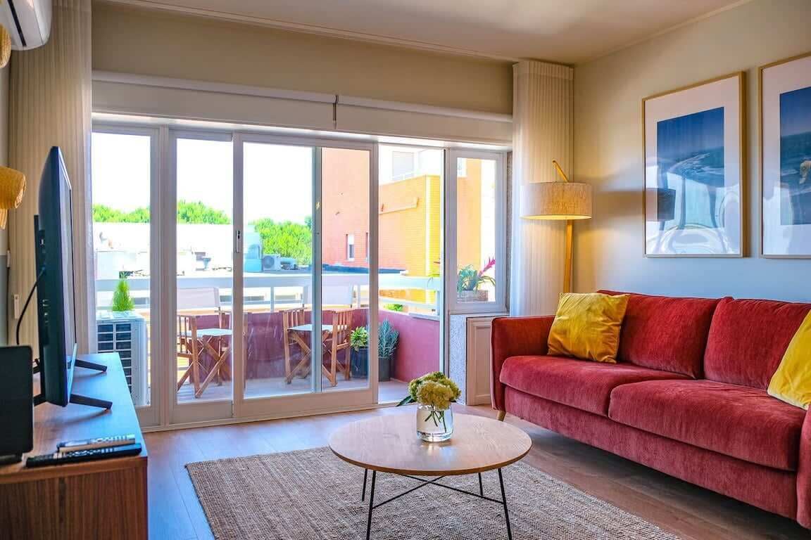 Un salón con un sofá rojo, una mesa de centro con un jarrón, un televisor sobre un mueble de madera y un ventanal corredero que da a un área exterior.