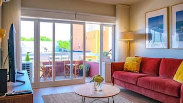 Un salón con un sofá rojo, una mesa de centro con un jarrón, un televisor sobre un mueble de madera y un ventanal corredero que da a un área exterior.