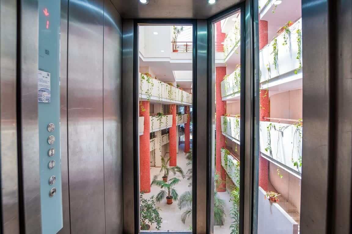 An interior view of an elevator with a glass door, revealing a multi-level atrium with plants and a red brick wall.