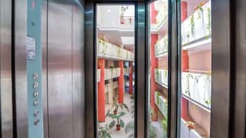 An interior view of an elevator with a glass door, revealing a multi-level atrium with plants and a red brick wall.