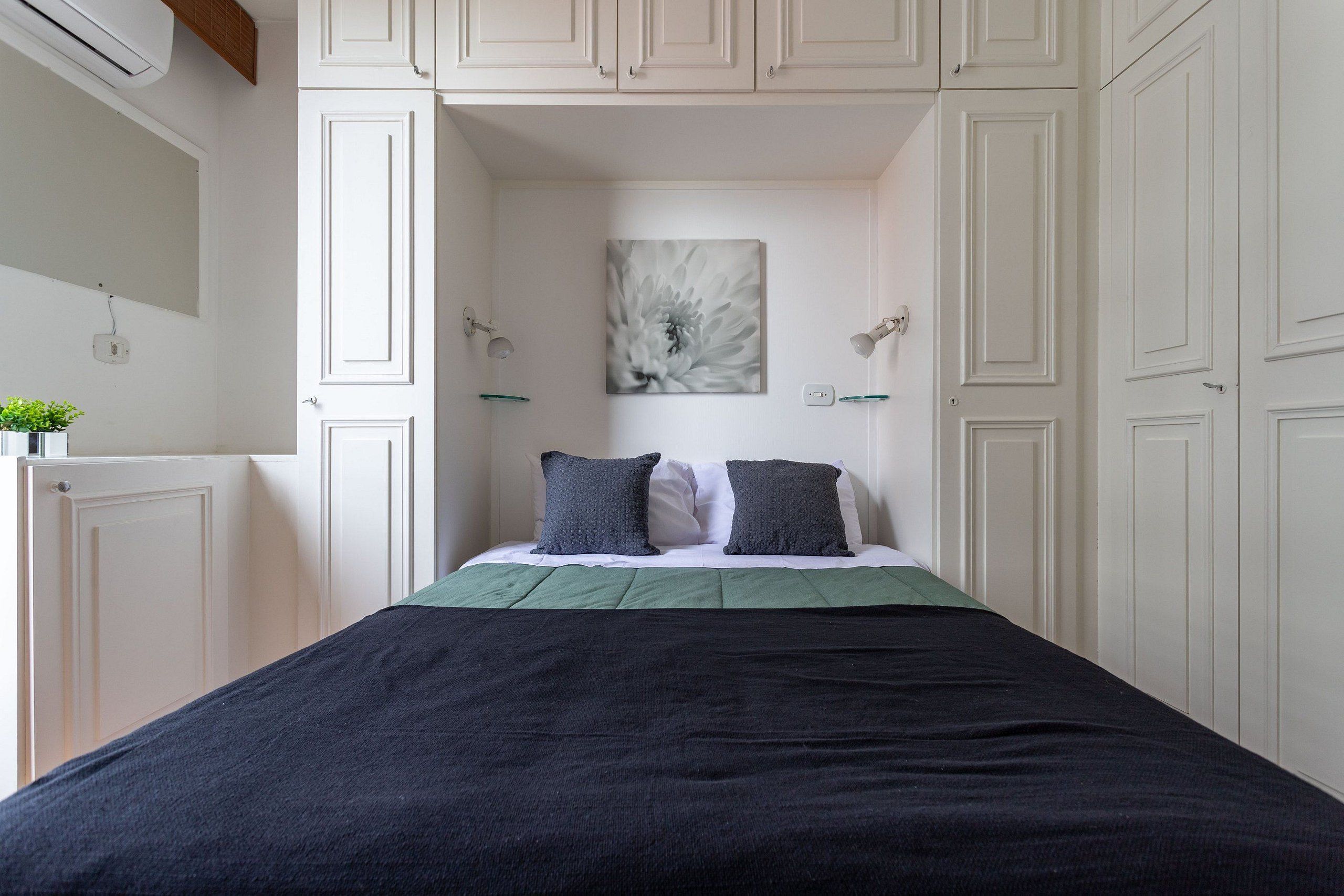 Quarto com uma cama, mesa de cabeceira, luminária na parede e um quadro.