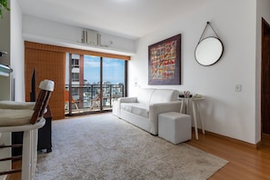 A modern living room with a white sofa, a small side table, and a large window offering a city view. - (1008) Apartamento com 1 Quarto e Vista para o Cristo (Rio de Janeiro)