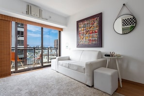 A modern living room with a white sofa, a glass table, and a city view through large windows. - (1008) Apartamento com 1 Quarto e Vista para o Cristo (Rio de Janeiro)