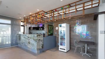 Uma área de bar moderna com bancada de mármore, geladeira repleta de bebidas e uma luminária pendente colorida.