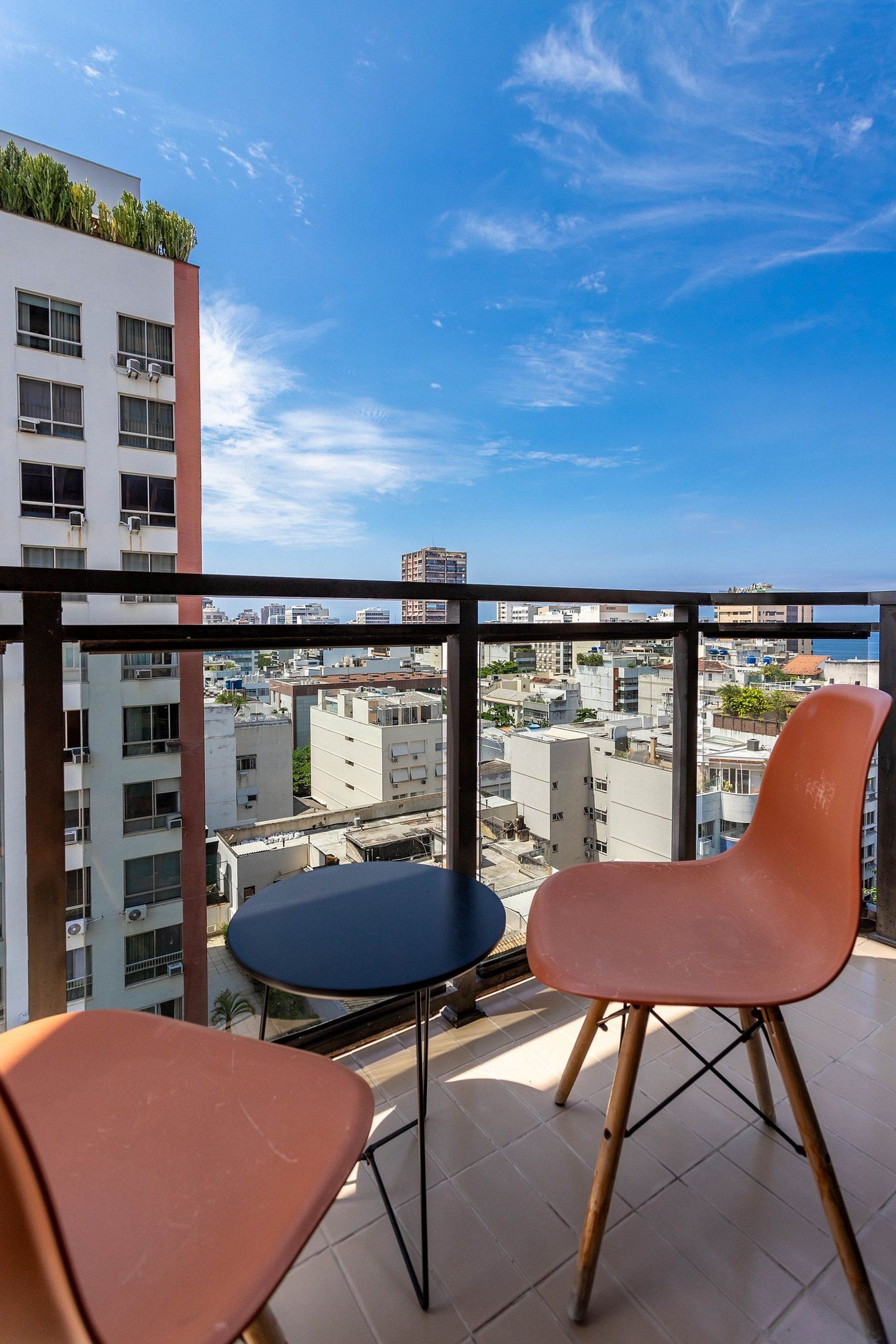Uma varanda com duas cadeiras e uma mesa pequena, com vista para um horizonte urbano com edifícios e céu claro.